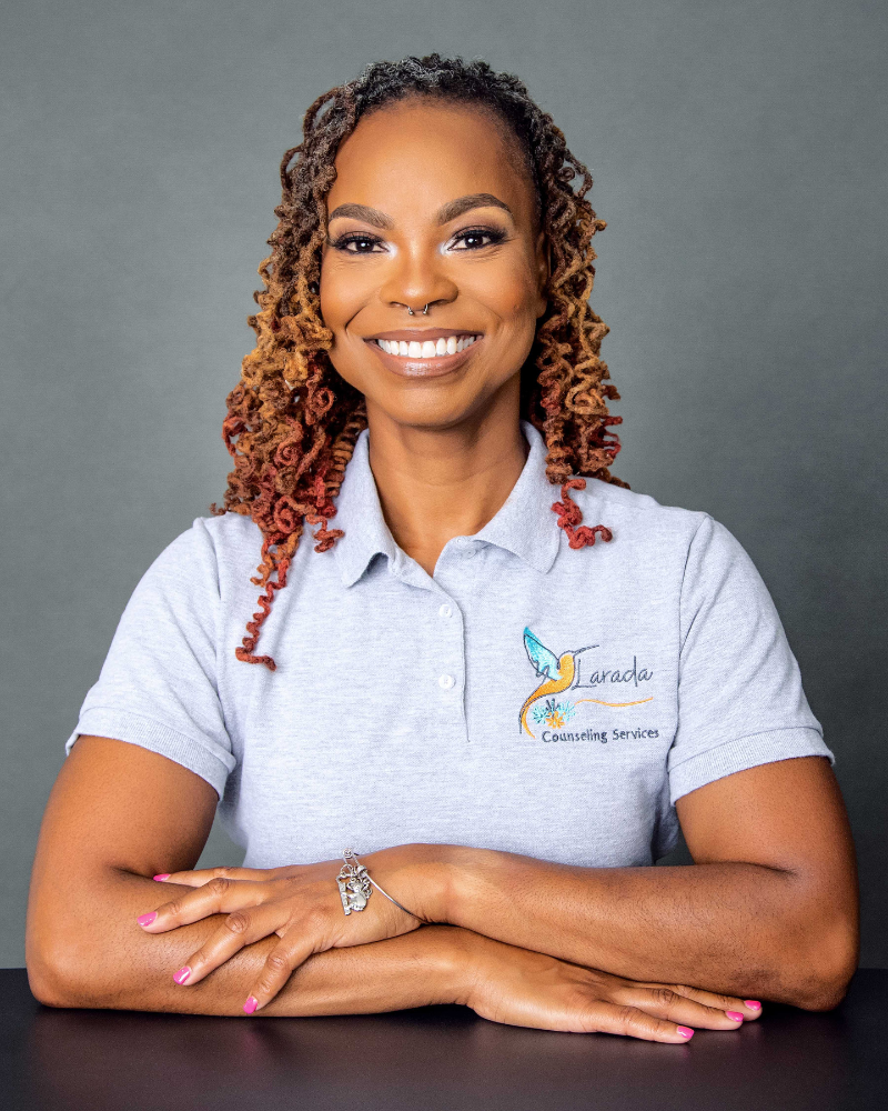 badia-khalid-manning-licensed-certified-professional-counselor Smiling woman with curly hair and nose ring, wearing a gray polo shirt with a counseling service logo, arms crossed on a table.